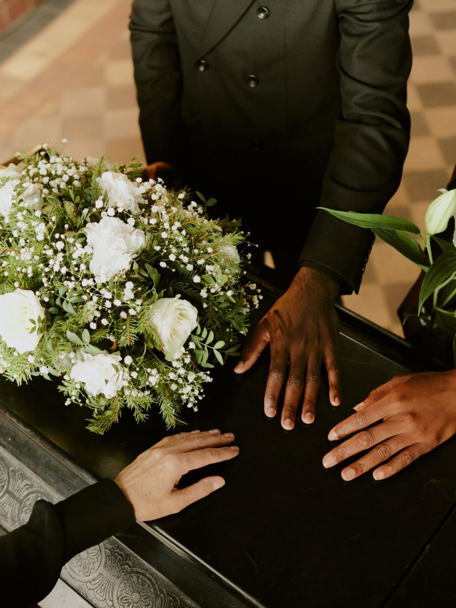 High angle shot of unrecognizable hands on black wooden coffin with white roses on top, pastor holding holy book
